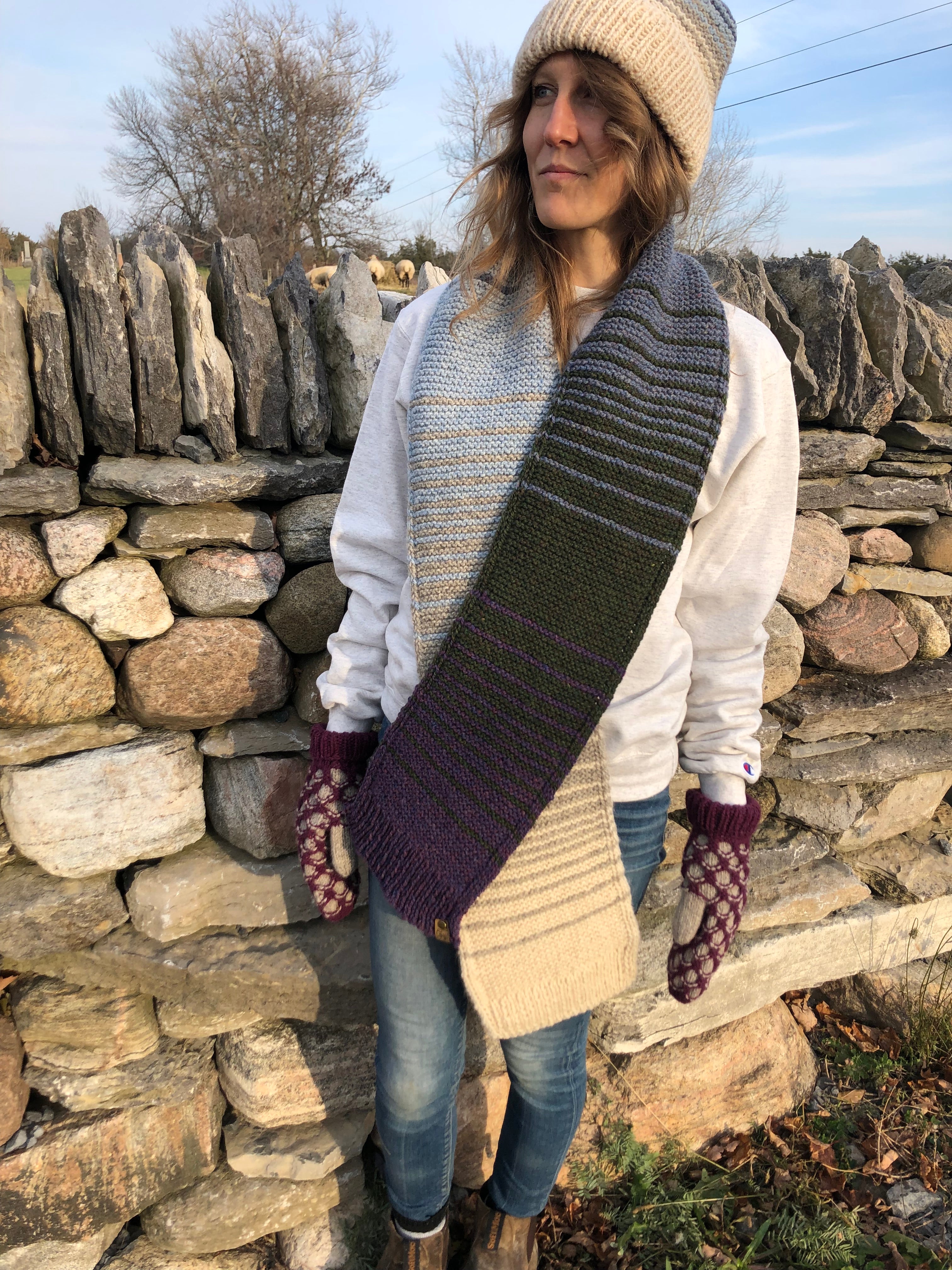 Blonde woman in front of a stone wall in Topsy Farms' Shoreline hand knit scarf and hat