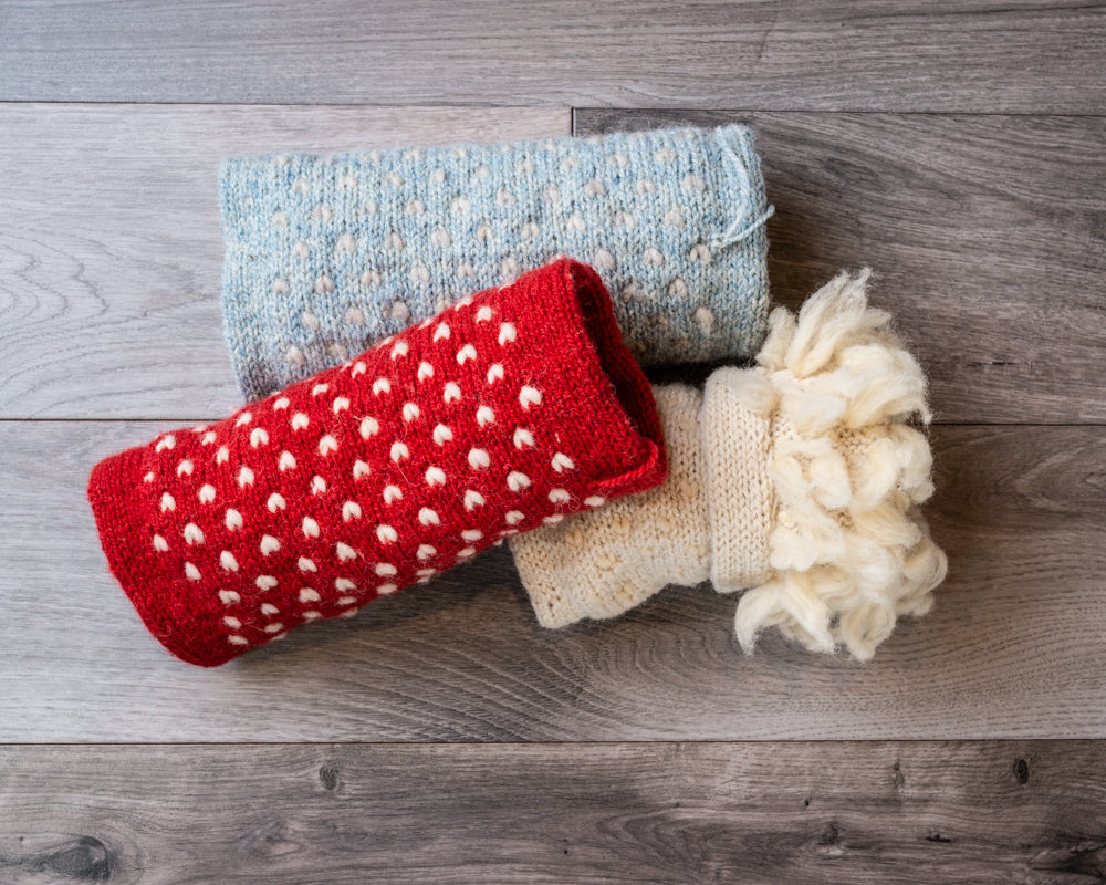 Red, light blue, and white hand-warming muffs with white accent Vs knitted throughout, on barnboard