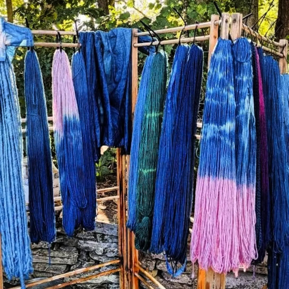 Multiple long skeins of tie-dye Topsy Farms' yarn hanging on wooden racks outdoors, showing blue, teal, pink, and purple color treatments against a natural forest background.