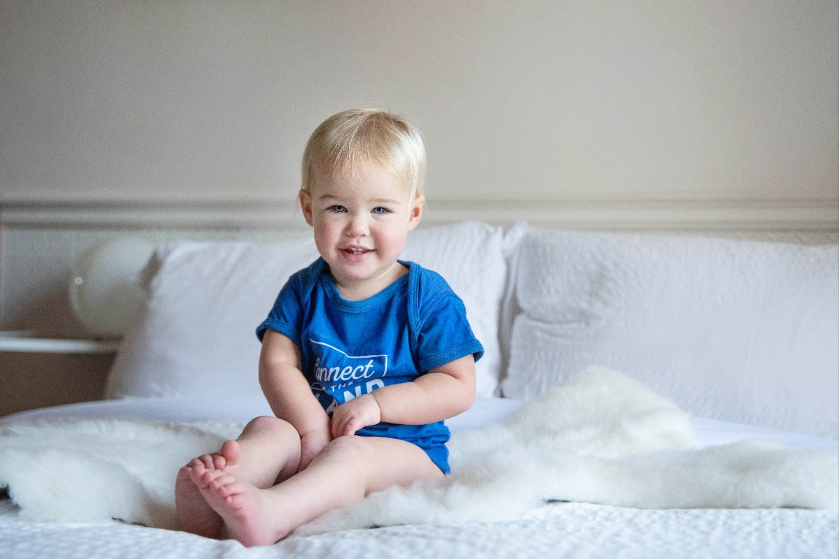 Blonde baby in Topsy Farms' connect to the land onesie sitting on a white lambskin on a white bed