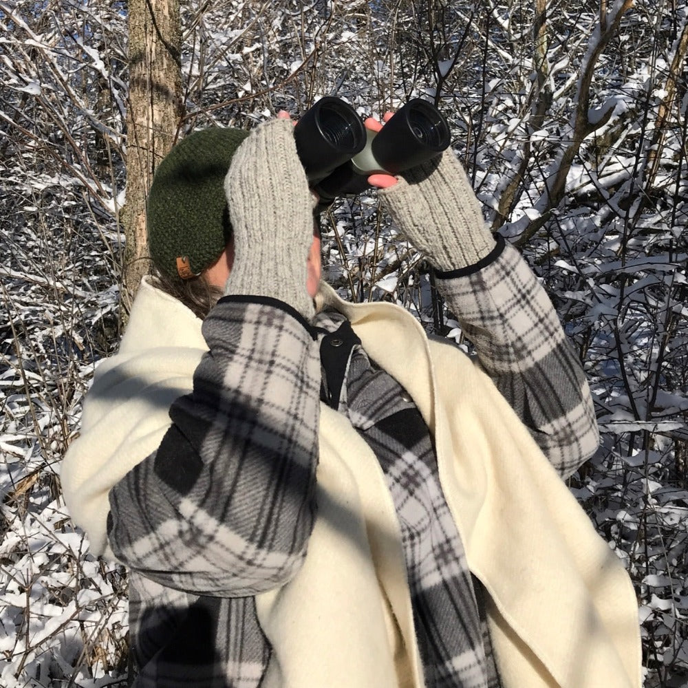 Person standing in snow covered woods, looking through binoculars, wearing Topsy Farms' wool wrap and fingerless gloves
