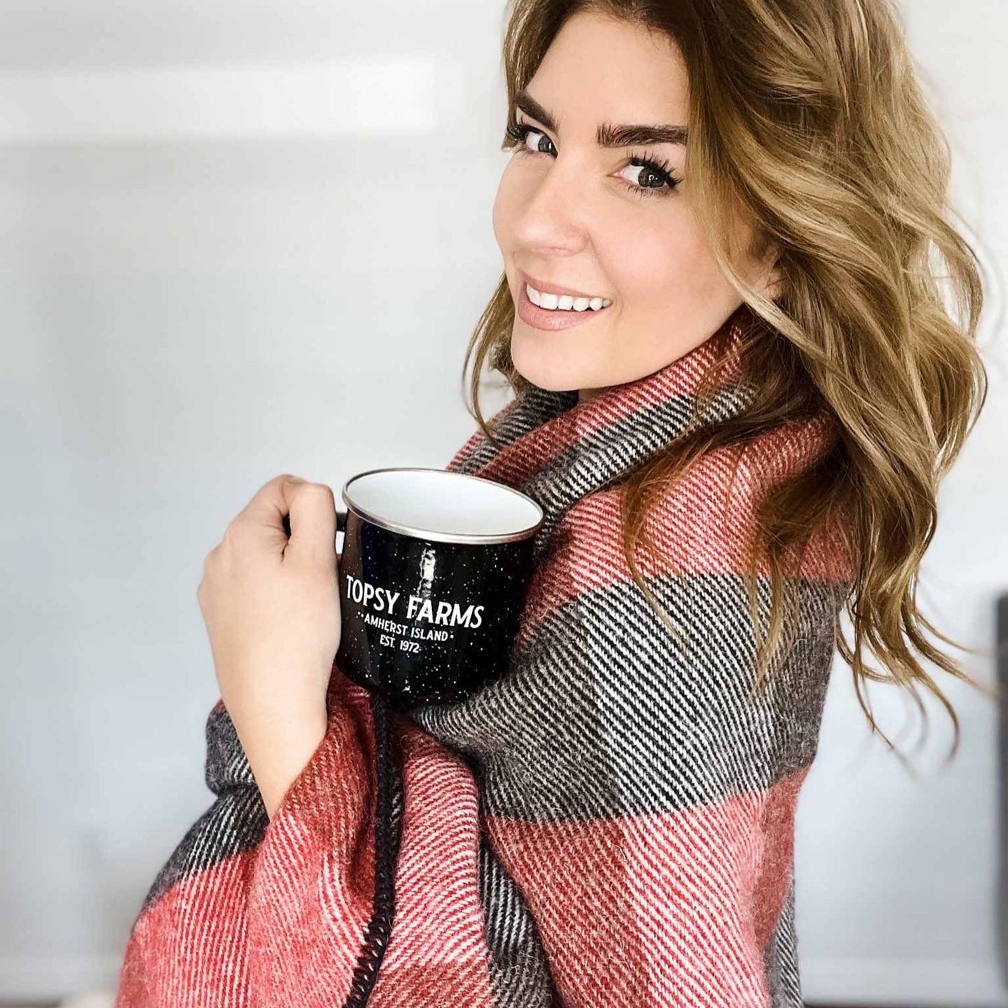 Blonde woman wrapped in Topsy Farms' red and black checkerboard wool blanket holding a Topsy mug