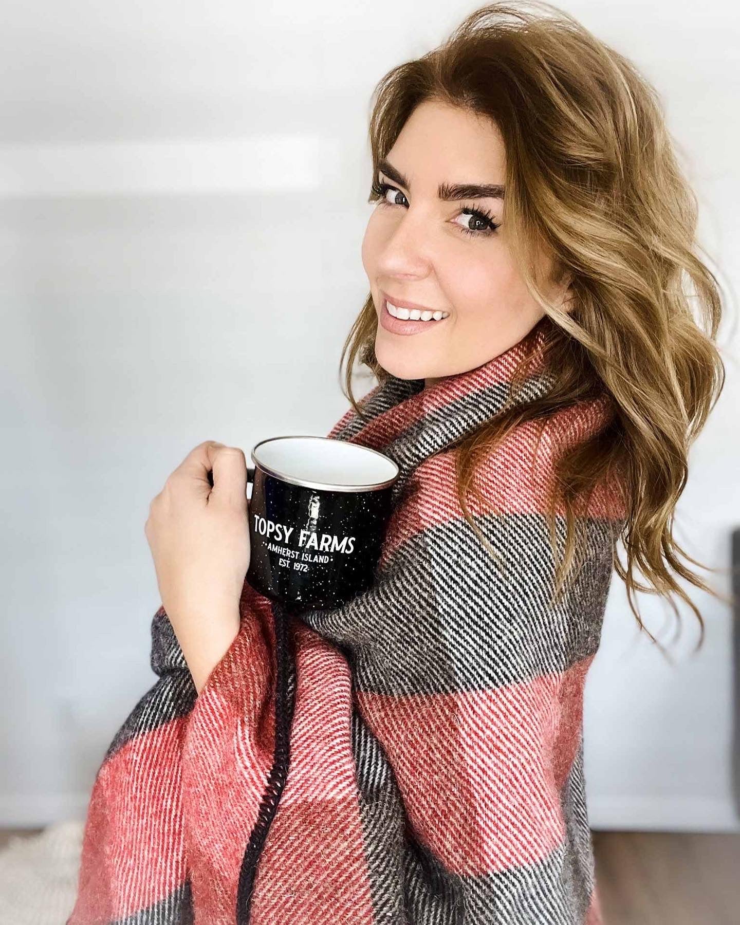 Blonde woman wrapped in Topsy Farms' red and black checkerboard wool blanket holding a Topsy mug