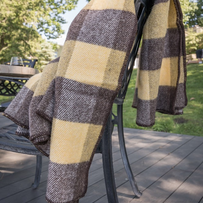 Topsy Farms' brown and gold checkerboard wool blanket, draped over a metal chair on a wooden deck with trees in the background
