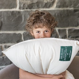 curly-haired child grinning and clutching Topsy Farms' Lighthouse Collection wool filled pillow, in front of a stone wall