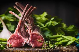 Two racks of lamb leaning against each other on a slate board, surrounded by green vegetables and heads of garlic