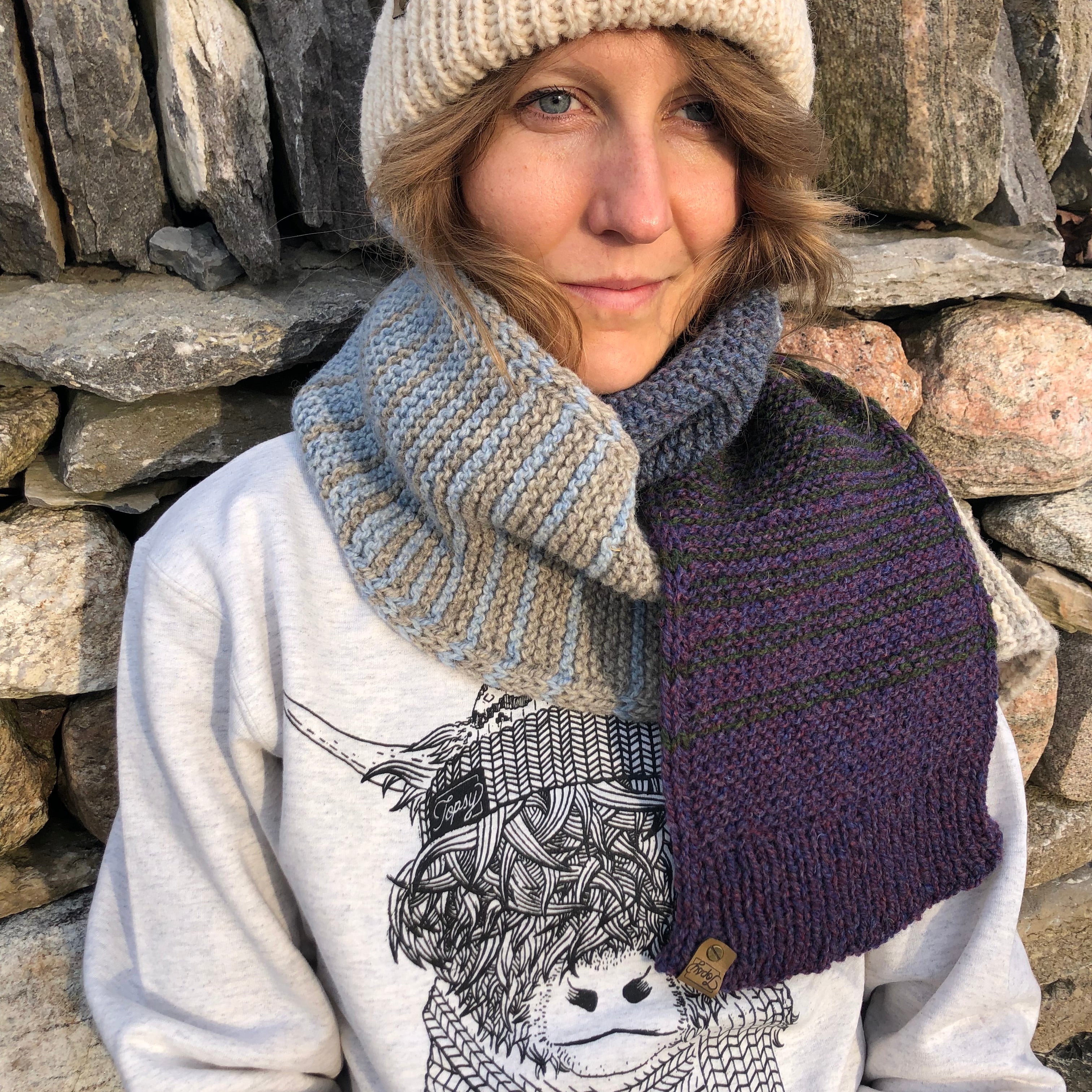 Blonde woman in front of a stone wall in Topsy Farms' Shoreline hand knit scarf and hat