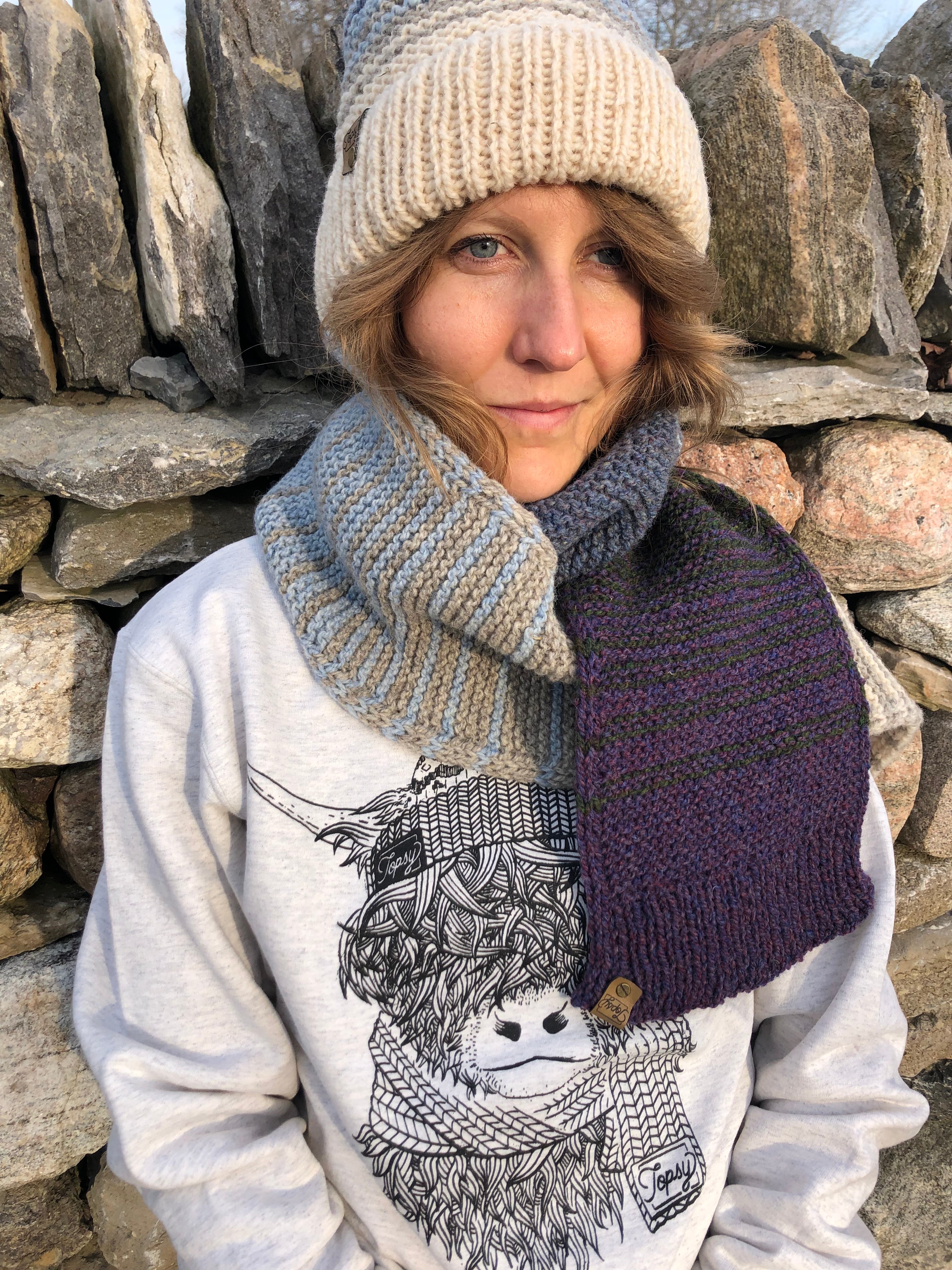 Blonde woman in front of a stone wall in Topsy Farms' Shoreline hand knit scarf and hat