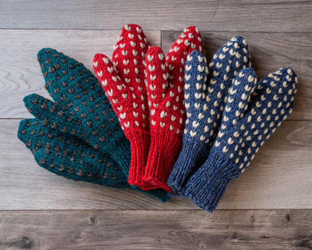 Green, red, and blue mittens with white accent Vs knitted throughout, on barnboard