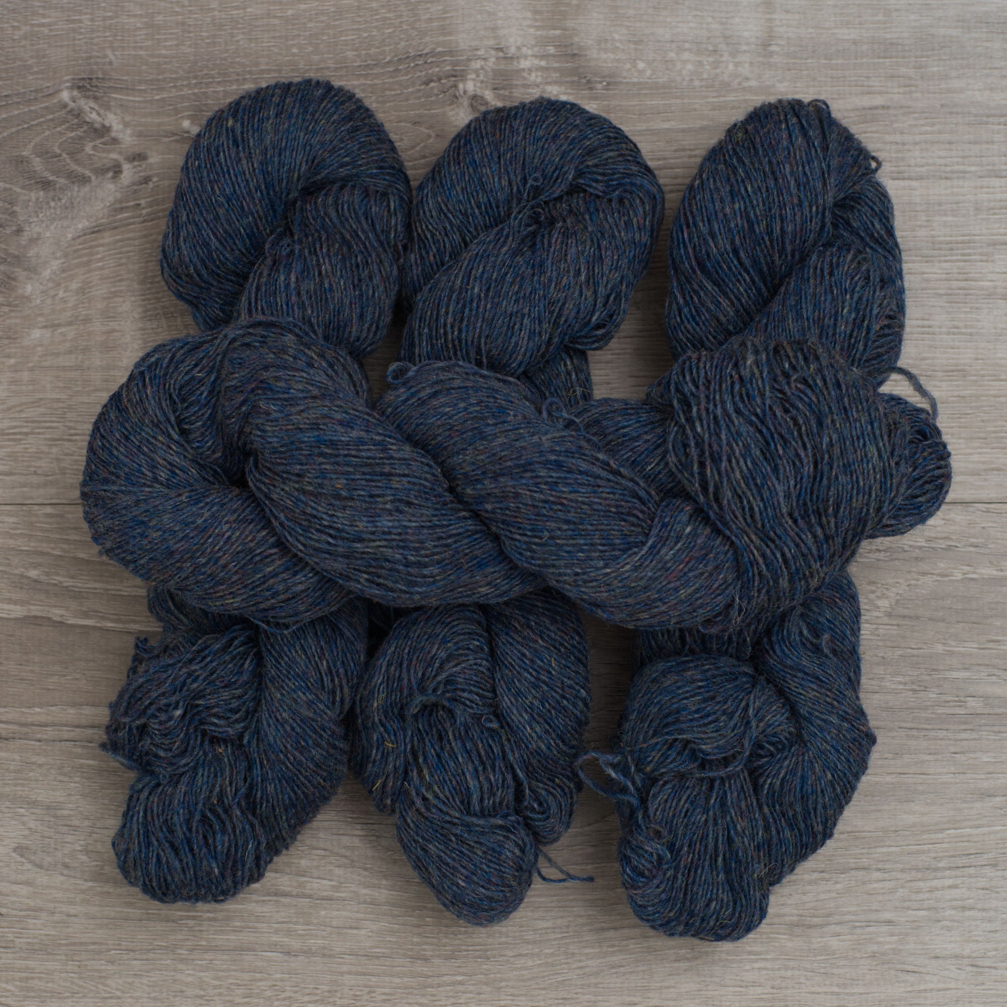 4 skeins of Topsy Farms' blue heather yarn in fingering weight, on a grey barn board background
